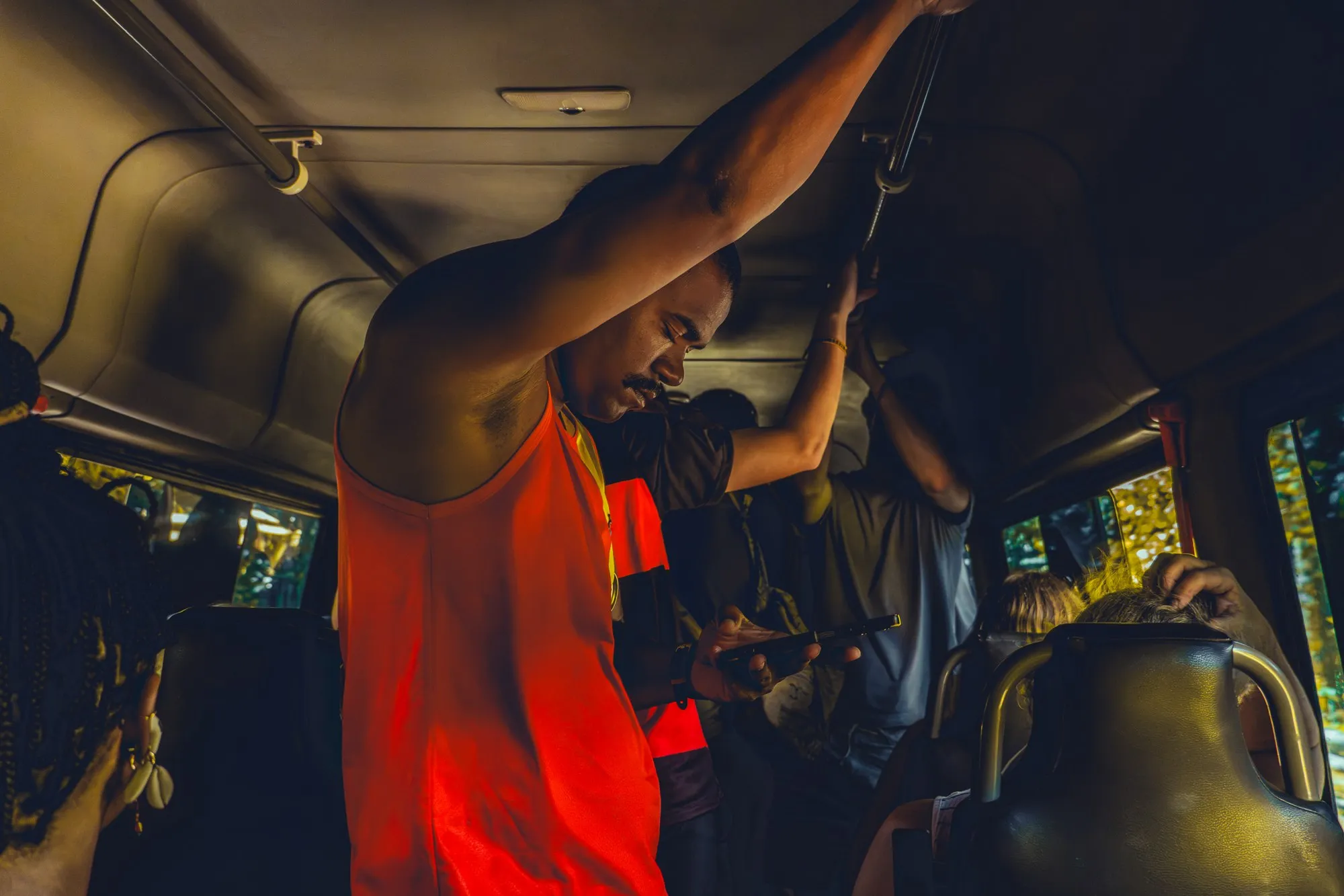Dans un bus, un homme en débardeur orange se tient à la barre du plafond.