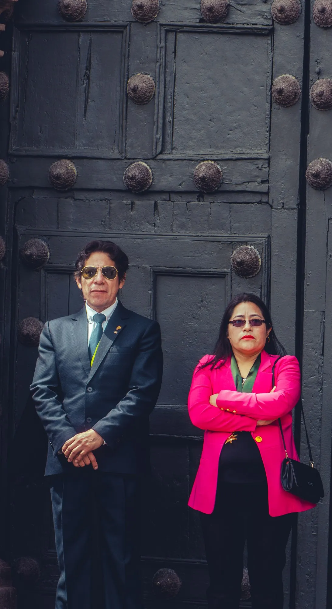 Un homme en costume et une femme en veste rose vif posent devant une lourde porte cloutée.