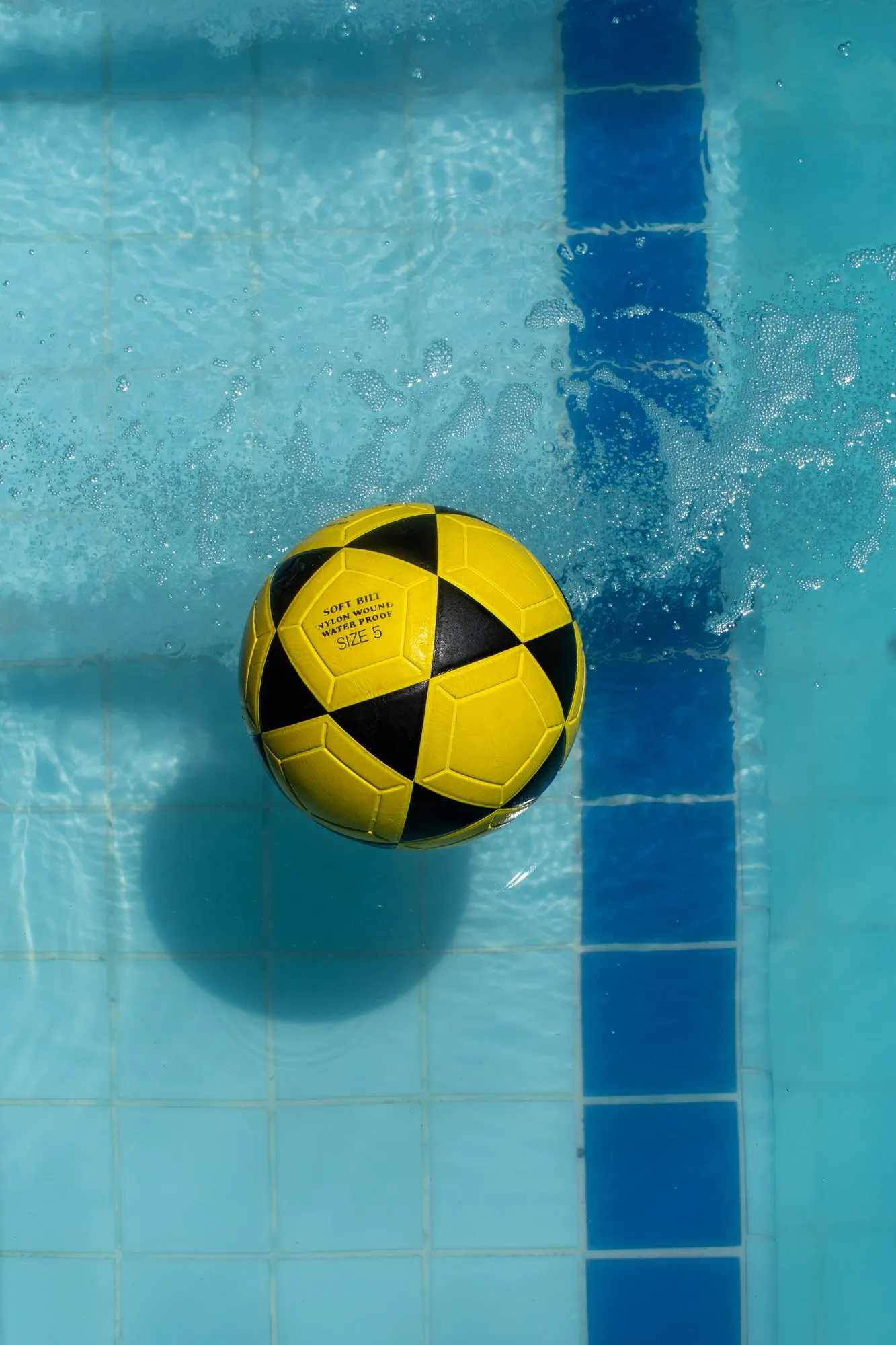 Un ballon jaune et noir flotte à la surface d'une piscine turquoise, près d'une ligne de carrelage bleu.