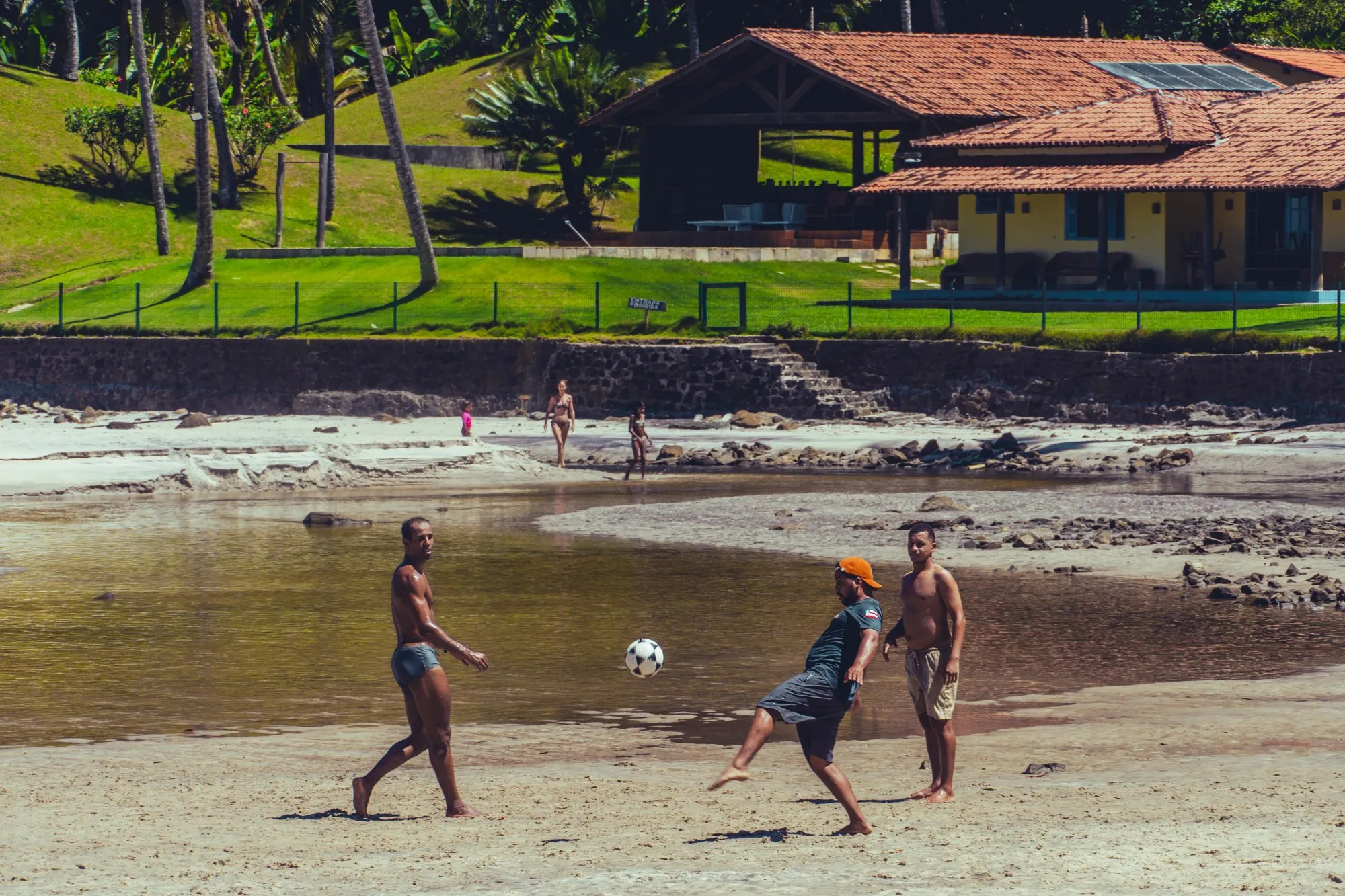 Trois hommes jouent au football sur une plage à marée basse, devant une maison au toit rouge.