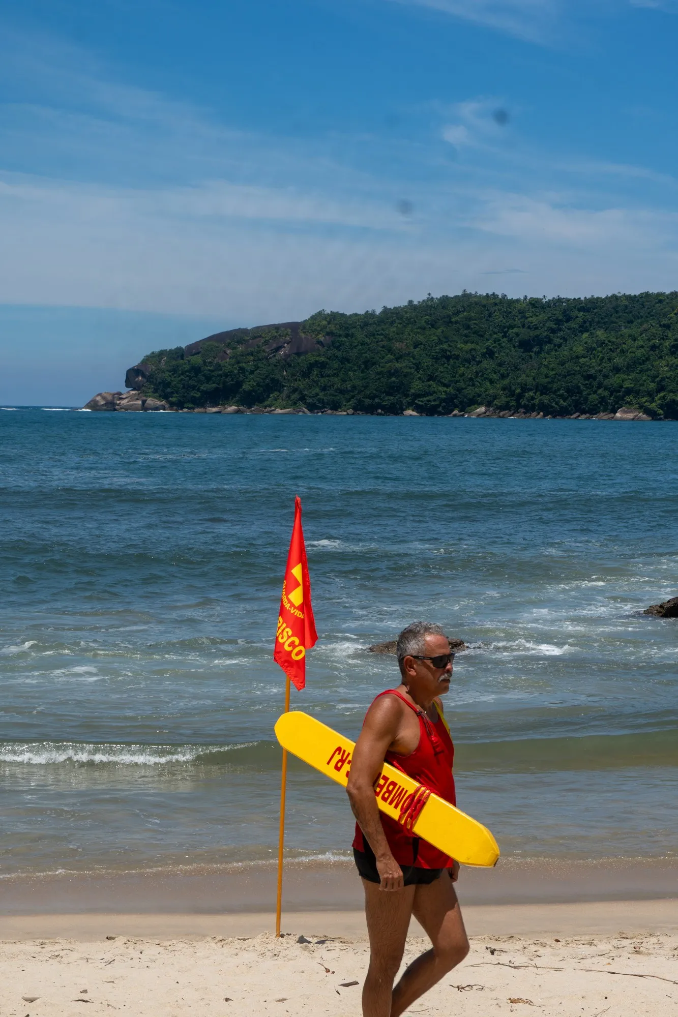 Un sauveteur en débardeur rouge marche sur le sable avec sa bouée jaune, devant un drapeau rouge planté face à la mer.