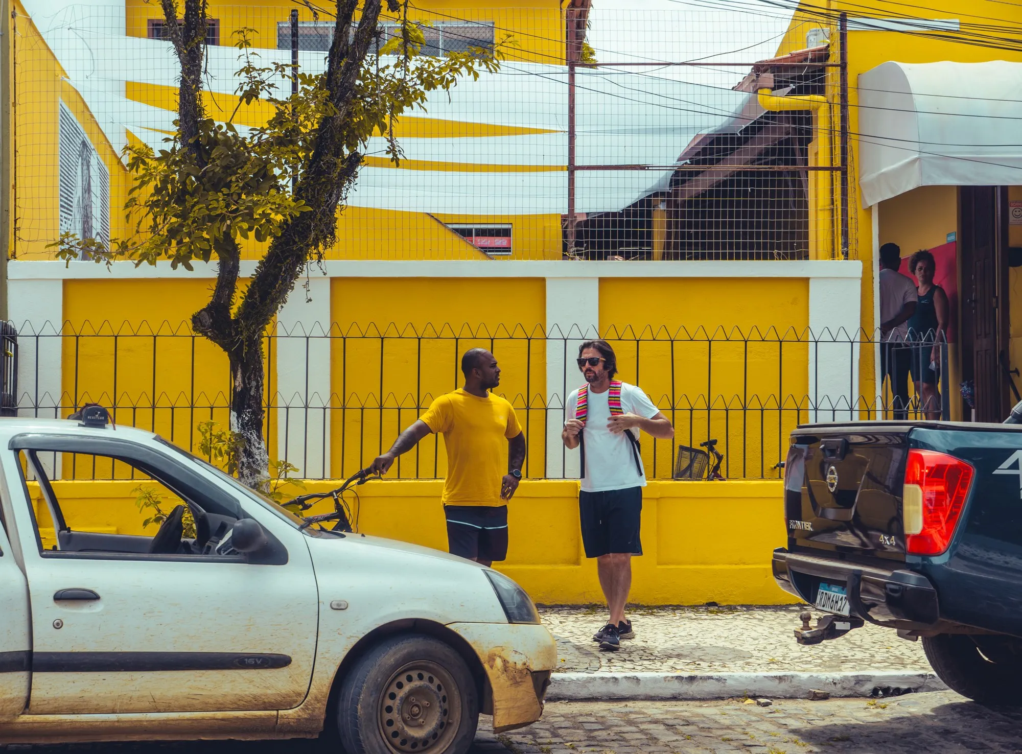 Deux hommes discutent devant un mur jaune, une voiture blanche garée au premier plan.