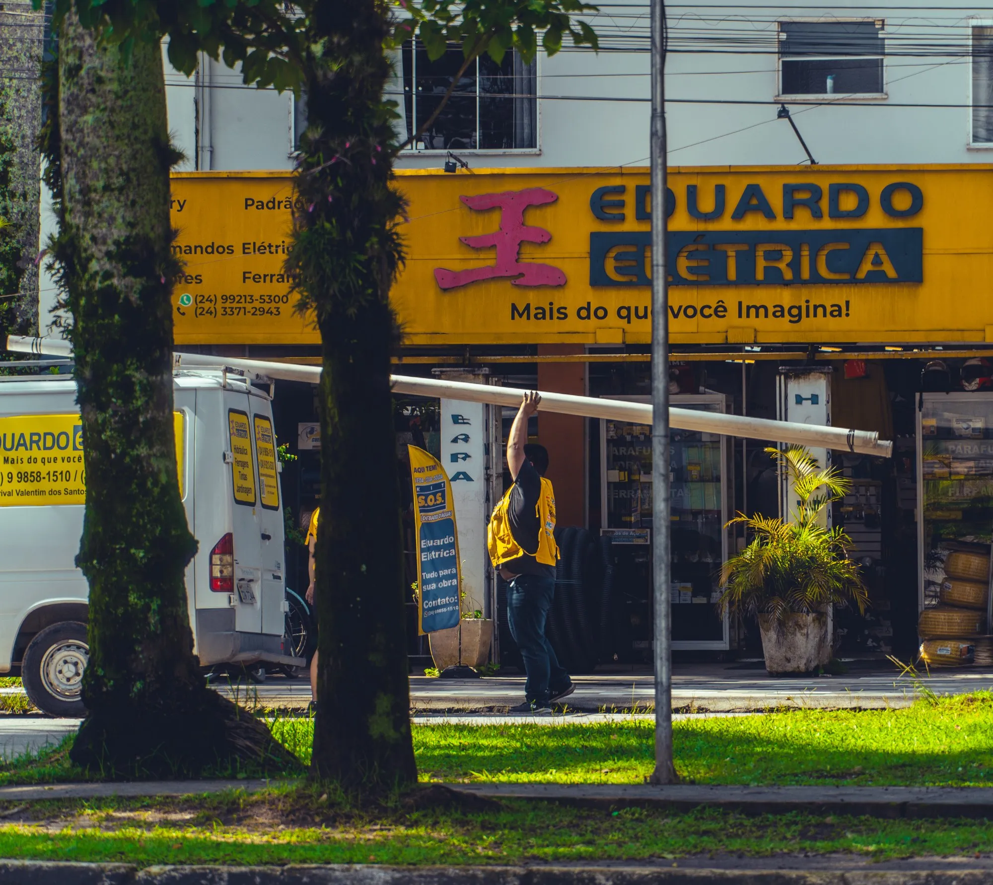 La façade jaune d'une boutique d'électricité, un homme en gilet jaune devant, entre deux troncs d'arbres.