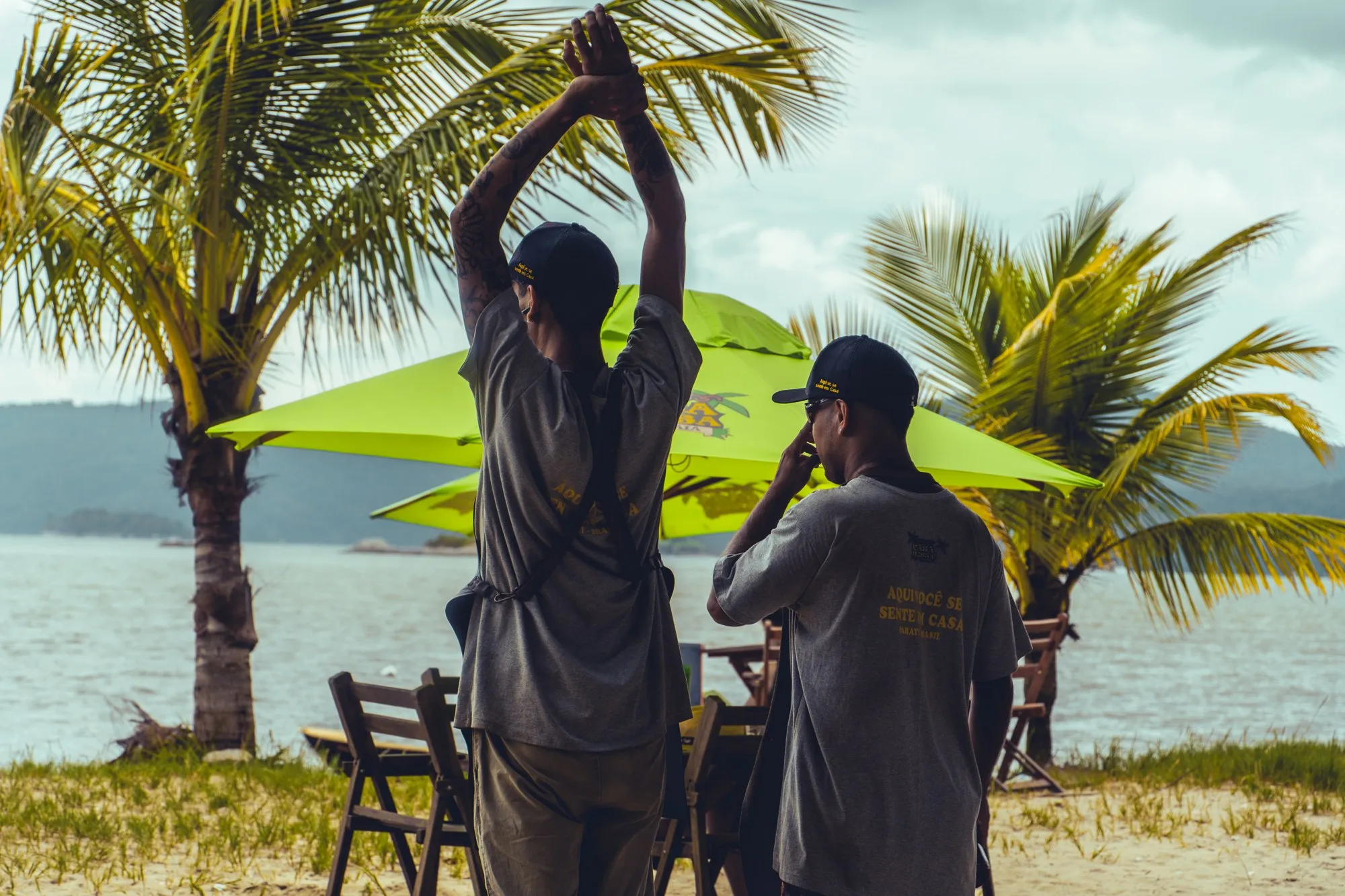 Deux hommes de dos devant des parasols verts et des palmiers, l'un s'étire les bras levés.