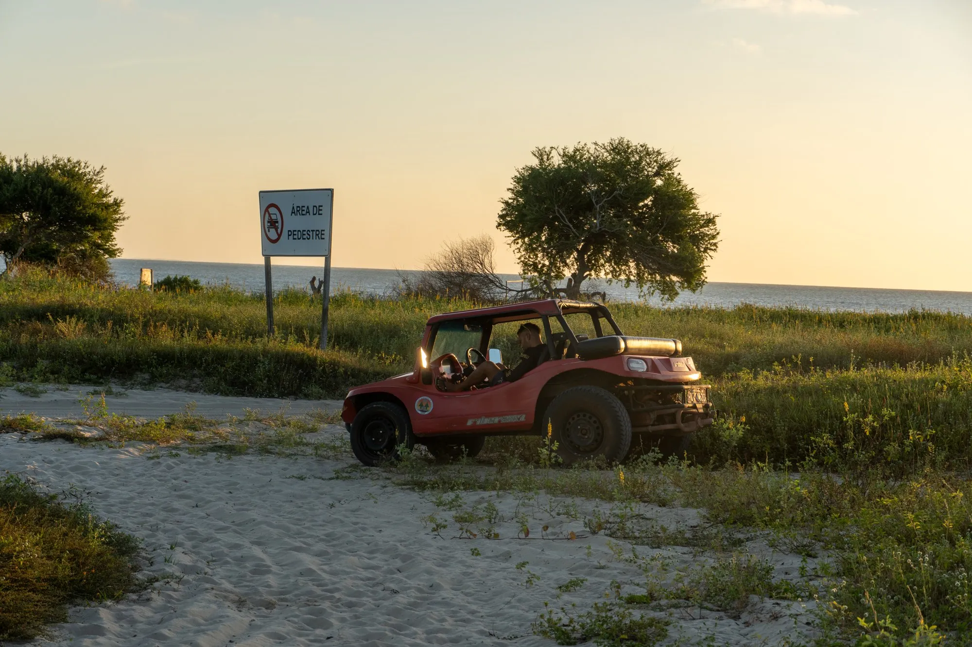 Un buggy rouge garé dans l'herbe au bord d'une piste de sable, au coucher du soleil.