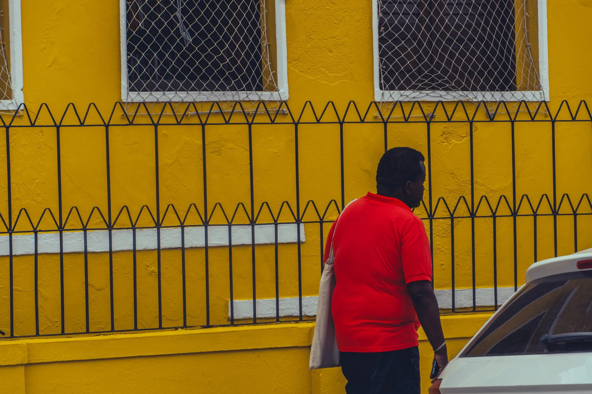 Un homme en t-shirt rouge longe une grille devant une façade jaune vif.