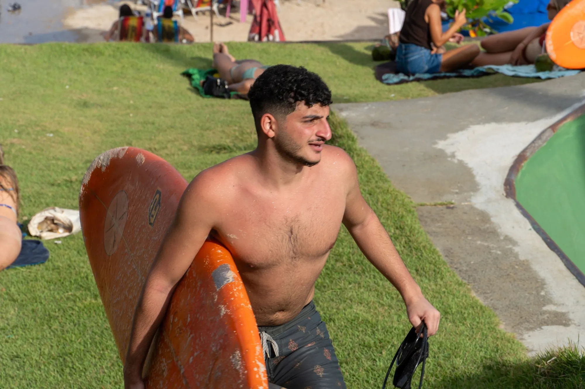 Un jeune surfeur torse nu sourit, sa planche orange sous le bras, sur une pelouse au bord de la plage.