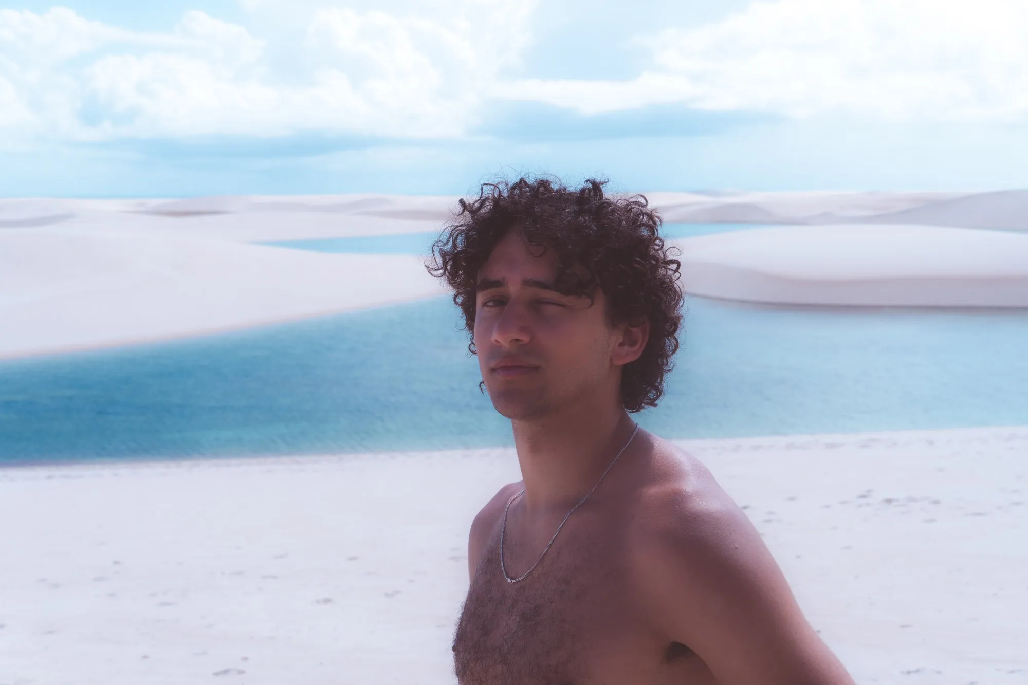 Un jeune homme aux cheveux bouclés, torse nu, se tient devant une lagune et les dunes.