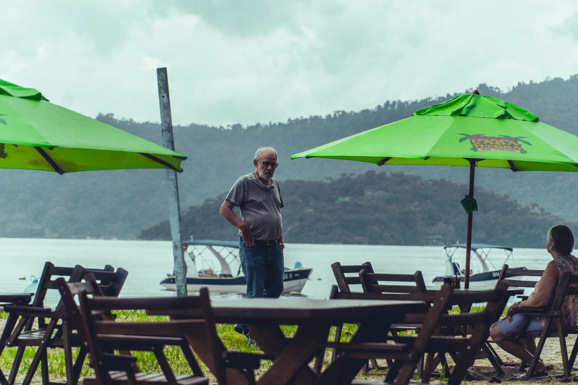 Un homme âgé debout entre des tables vides et des parasols verts au bord de l'eau.
