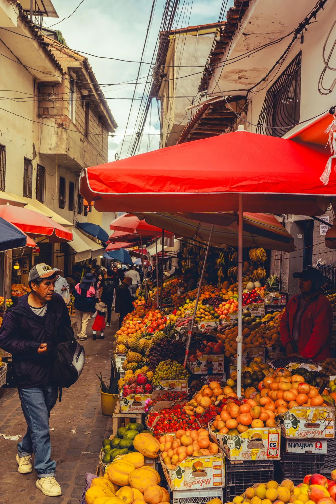 Une ruelle de marché, un grand parasol rouge au-dessus d'un étal de fruits orange.