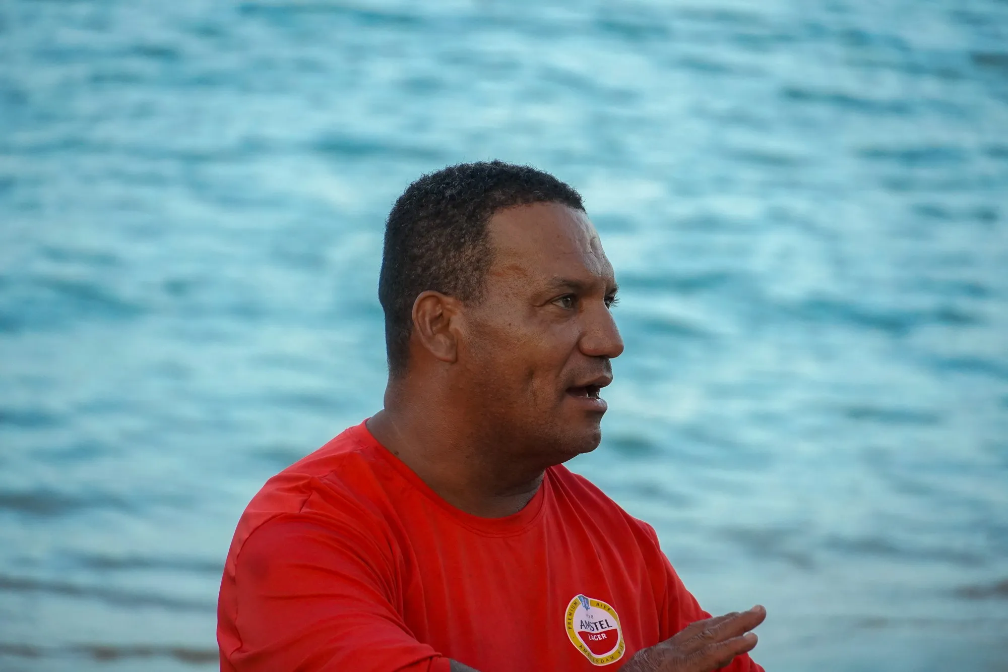 Portrait serré d'un homme en t-shirt rouge qui parle, devant l'eau bleue floue.