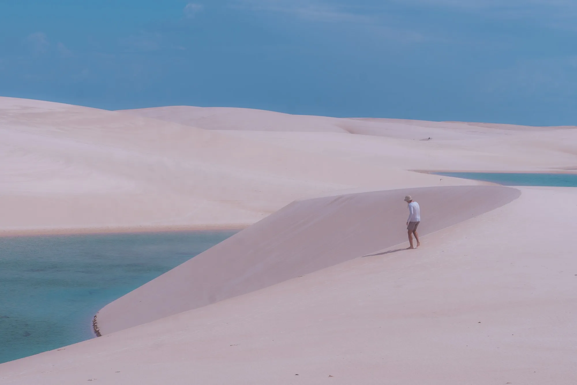 Une petite silhouette descend la pente d'une grande dune qui plonge dans l'eau bleue.