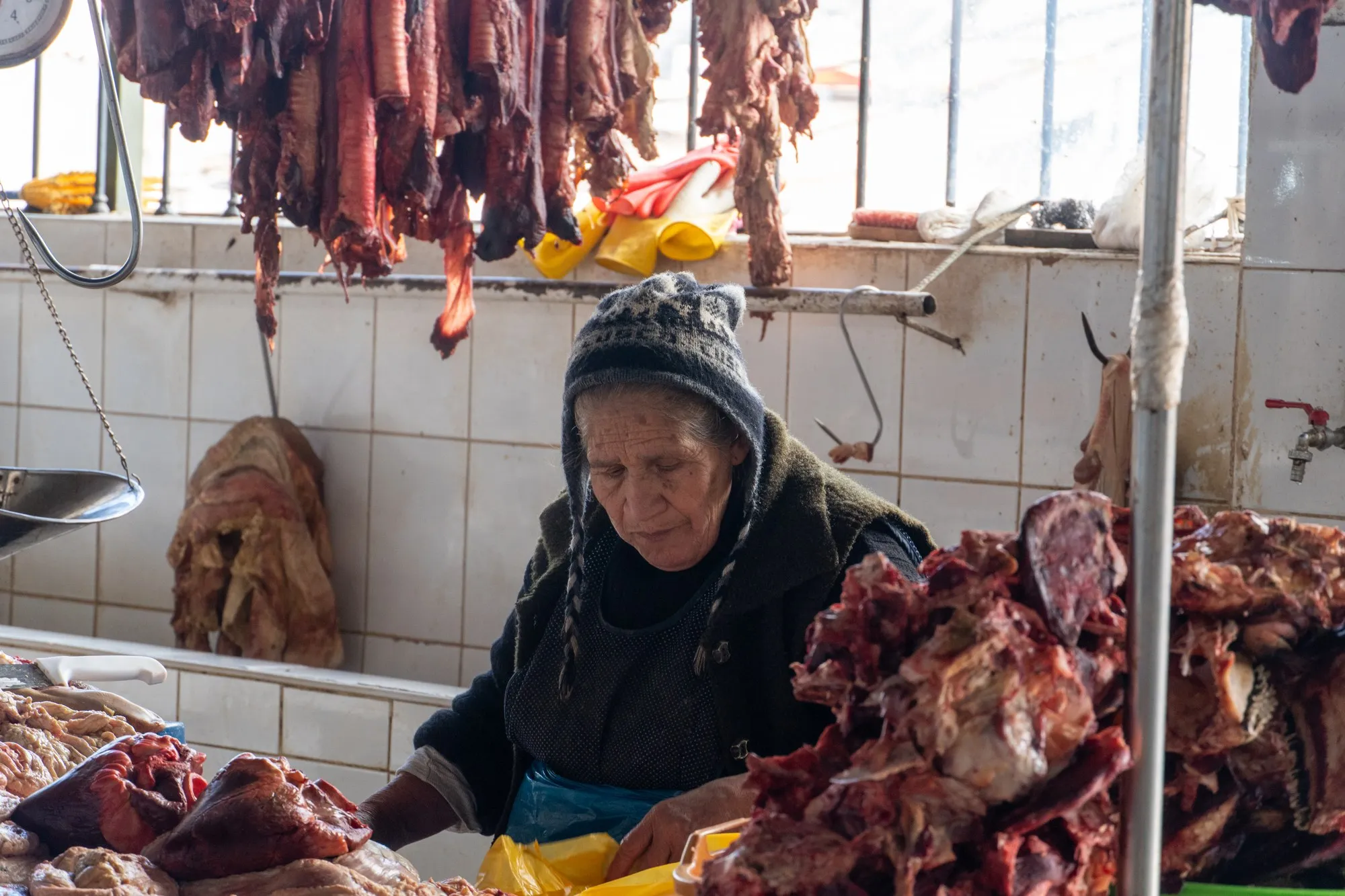 Une femme âgée en bonnet de laine découpe de la viande sous des pièces suspendues.