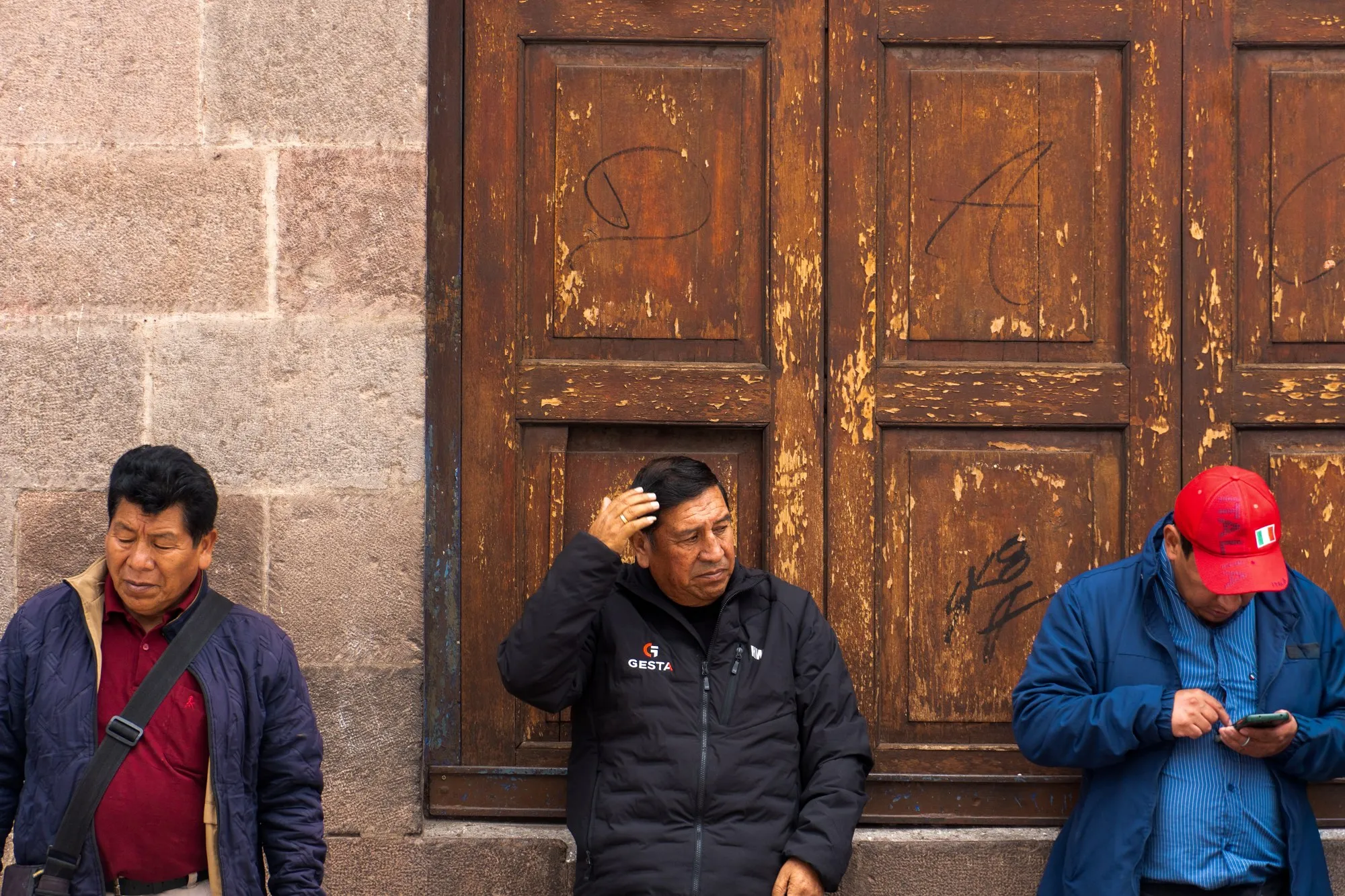 Trois hommes adossés à une vieille porte en bois, l'un d'eux regarde son téléphone.