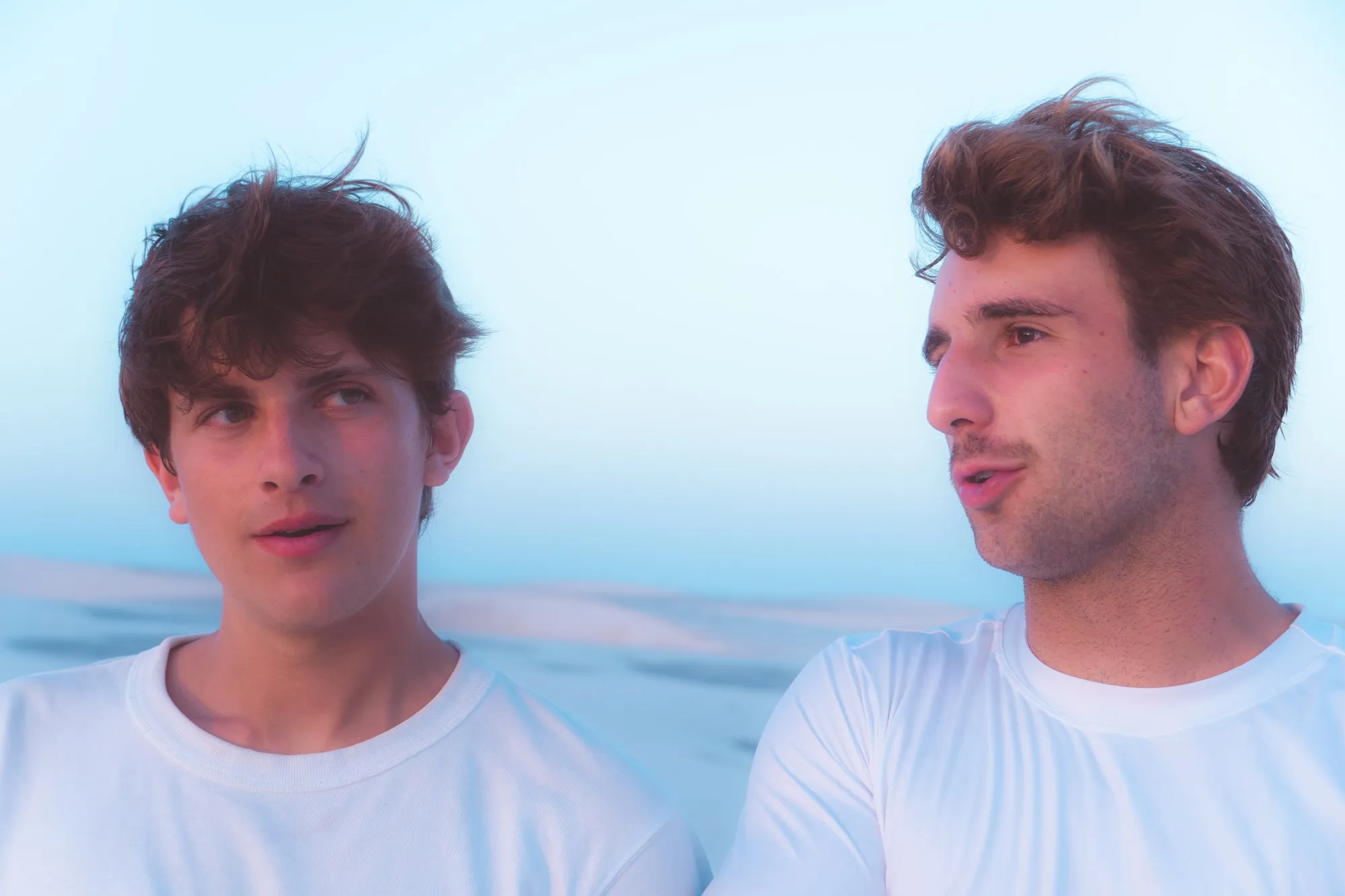 Portrait de deux jeunes hommes en t-shirt blanc devant un ciel pâle, le regard au loin.